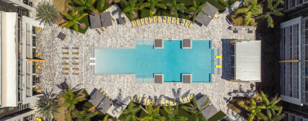 Aerial view of a large geometric resort pool surrounded by stone pavers, cabanas, and lounge chairs between two buildings.