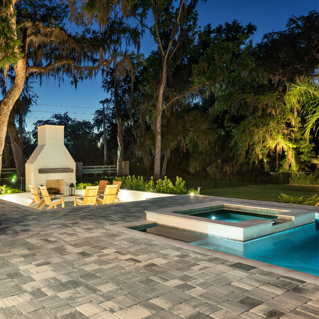 A raised spa glows turquoise at dusk beside a pool, with an outdoor fireplace and lounge seating nearby.