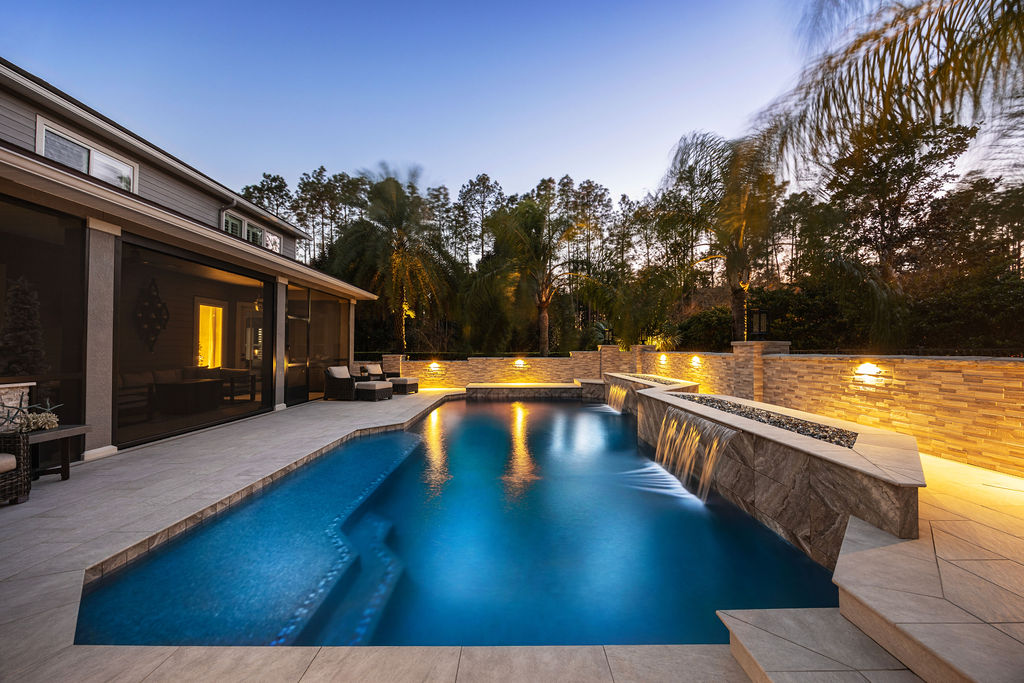 A dusk view of a modern rectangular pool with a raised stone wall waterfall feature and illuminated backyard landscaping.