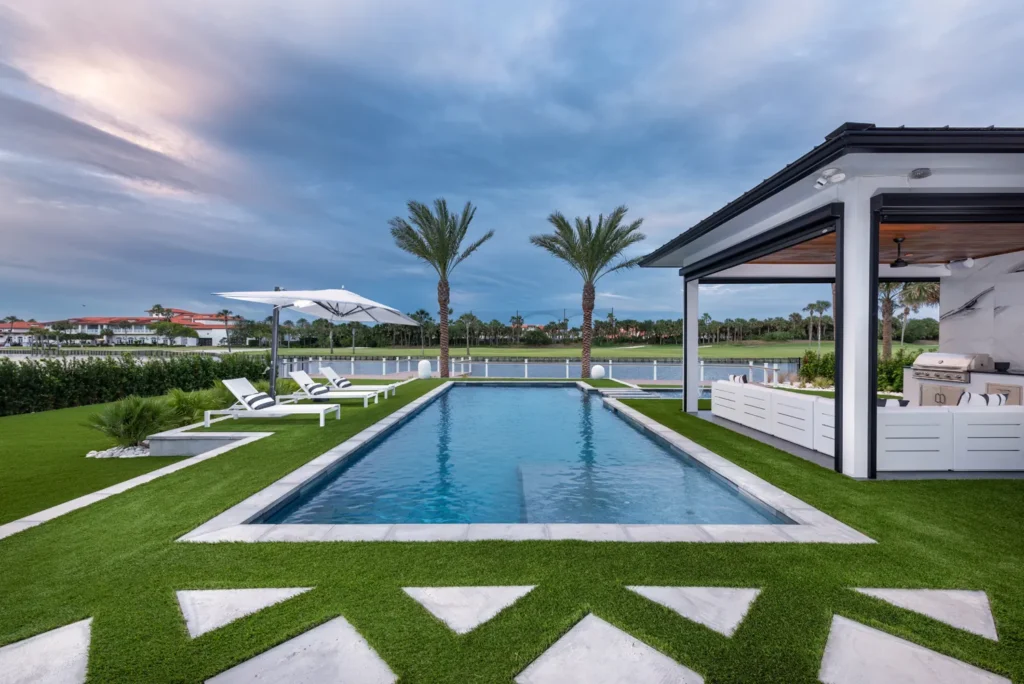 A rectangular pool with a covered outdoor kitchen sits beside a canal at dusk, framed by palm trees and artificial turf.