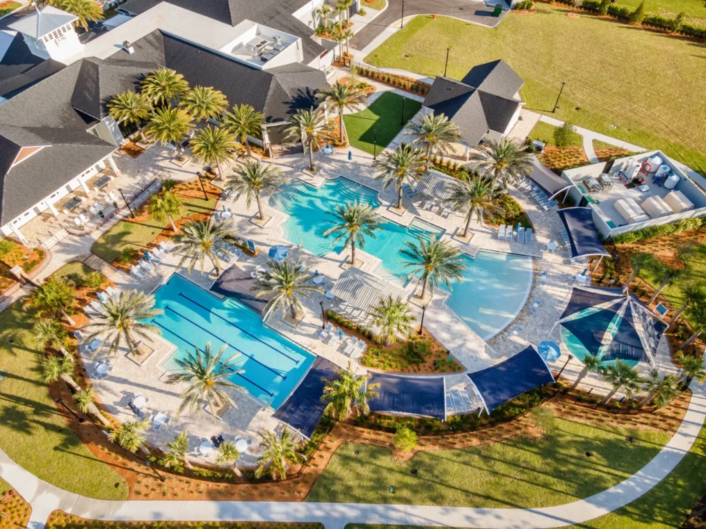 Aerial view of a resort-style community pool complex with a freeform main pool and a separate lap pool surrounded by palm trees and shade sails.