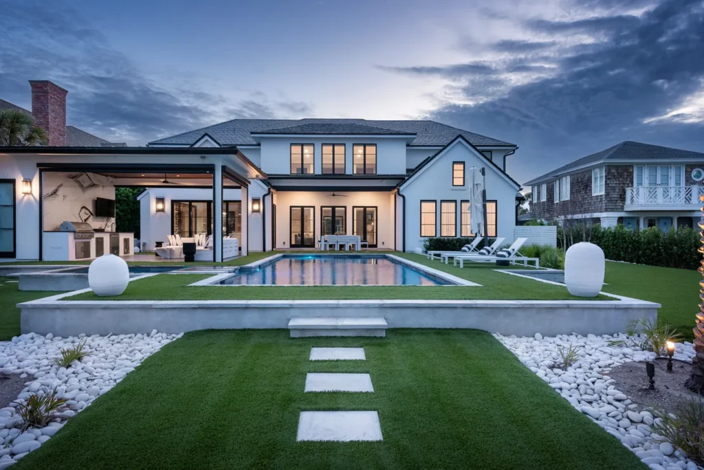 A rectangular backyard pool at dusk in front of a large two-story home with a covered outdoor kitchen and lush turf landscaping.