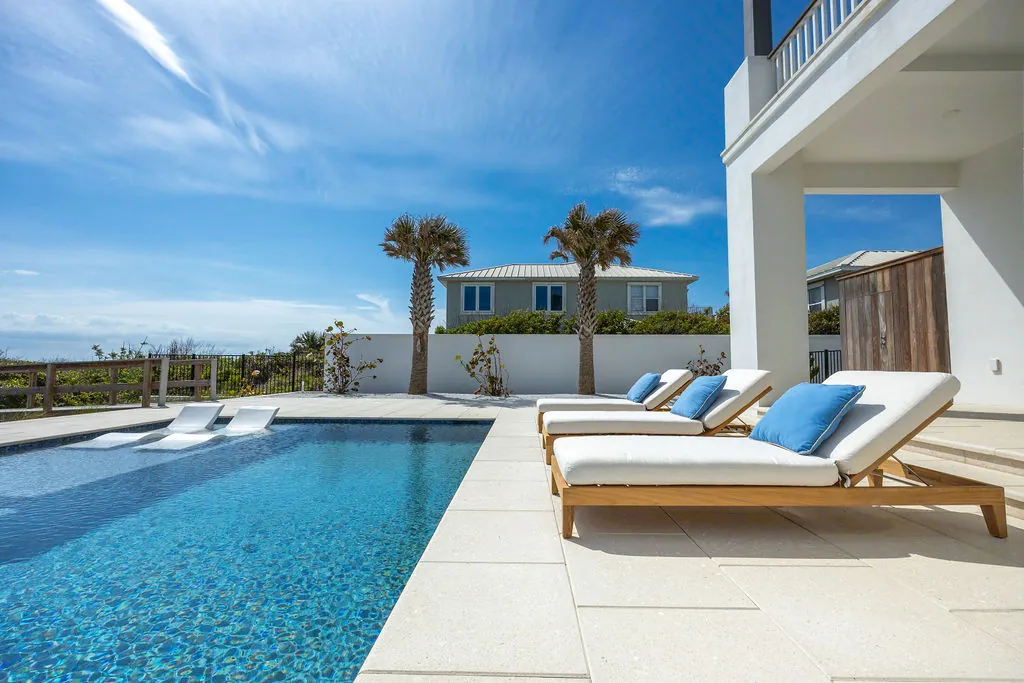 Lounge chairs on a stone patio beside a pool with a coastal home visible beyond palm trees.
