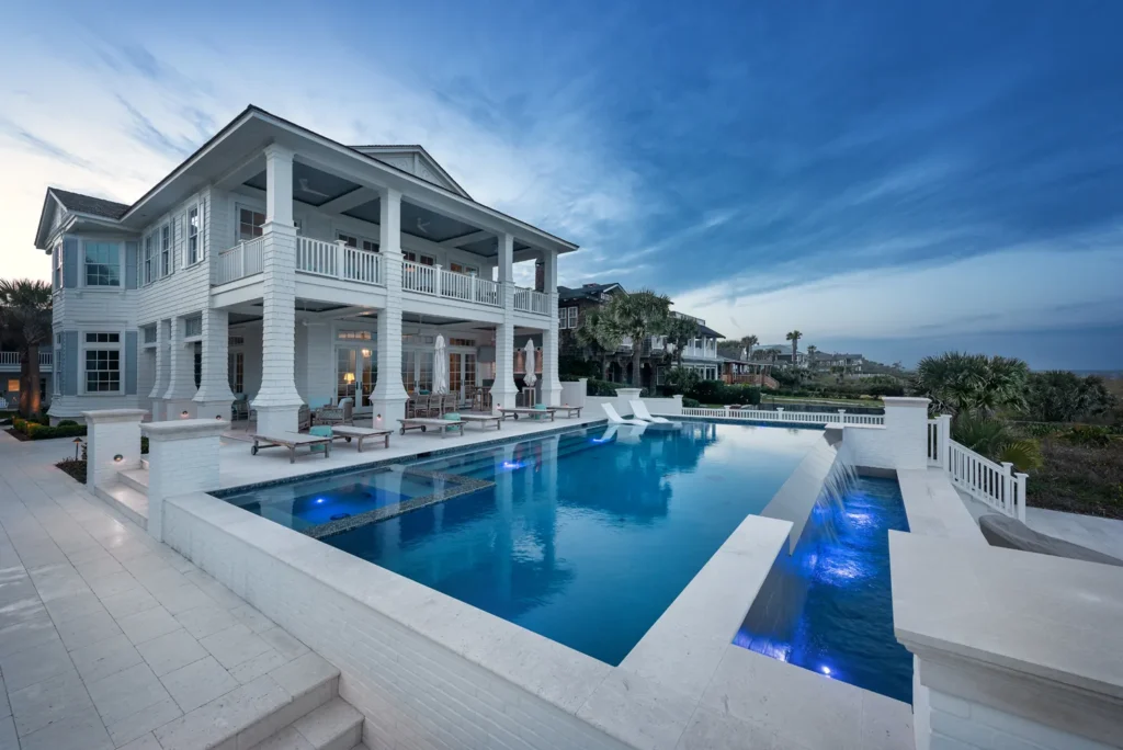 A large rectangular infinity-edge pool with a raised spa and waterfall spillover in front of a grand coastal home at dusk.