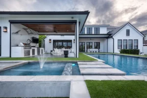 A long rectangular pool with a fountain jet and stone stepping pads spans the backyard of a modern white two-story home at dusk.