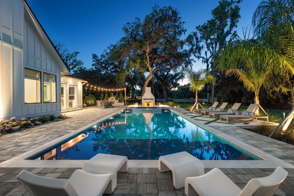 Rectangular pool at dusk with a stone fireplace at the far end and lounge chairs on a paver patio.