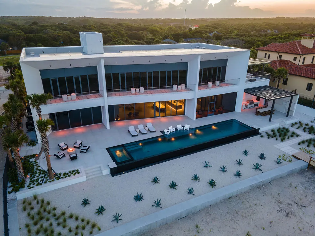 An elevated modern home at dusk with a narrow lap pool, raised spa, and fire bowls along a stone terrace.