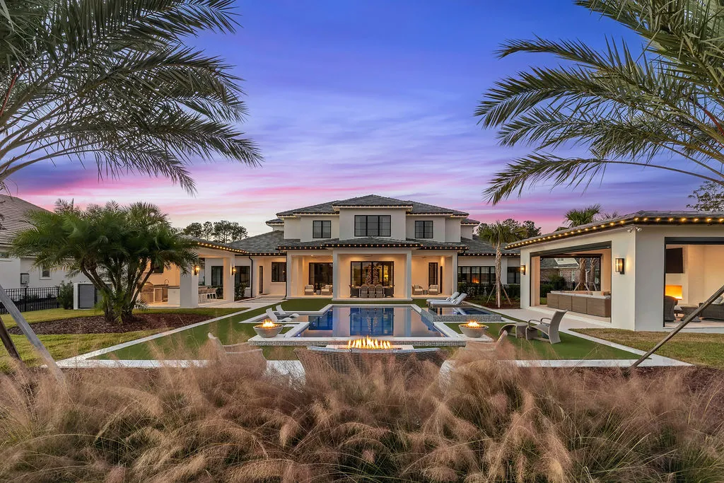 A large modern home at dusk with a rectangular pool, fire bowls, and a linear fire pit on the patio.