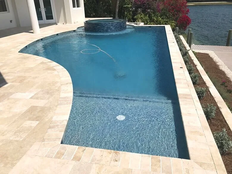 A curved infinity-edge pool with a raised tiled spa overlooks a canal behind a travertine-paved patio.