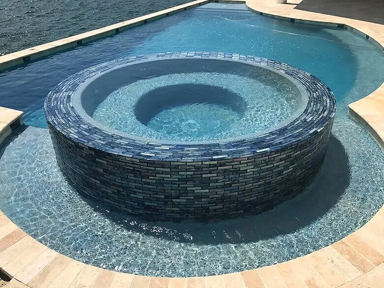 A round raised spa covered in blue glass tile spills over into a surrounding pool with travertine decking.