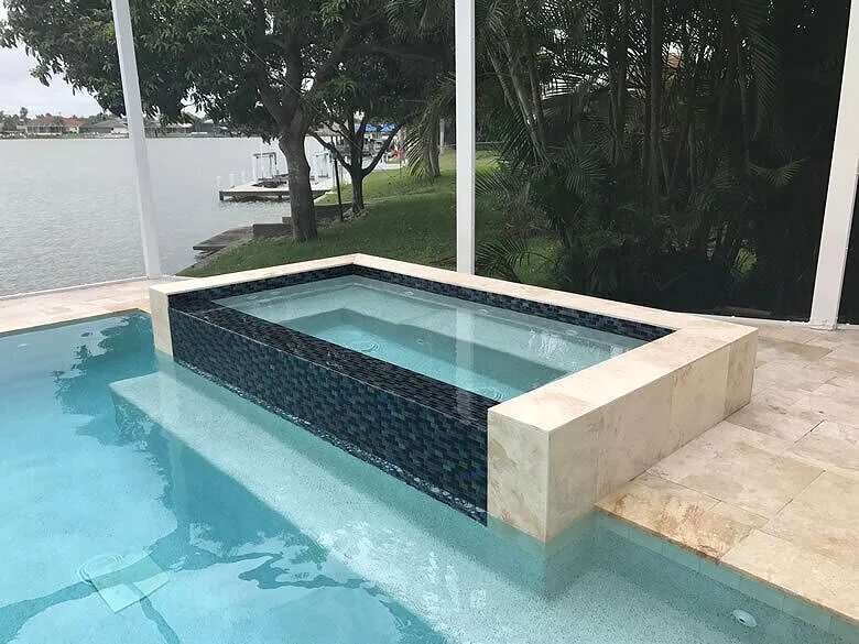 A raised spa with dark mosaic tile sits beside a pool overlooking a canal, enclosed by a screened lanai.