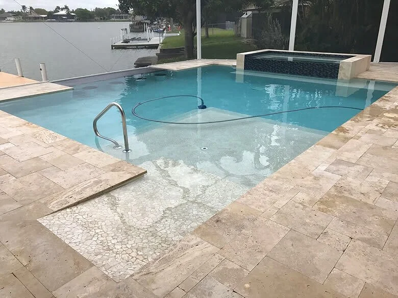A rectangular pool with a raised tiled spa sits on a travertine deck overlooking a canal.