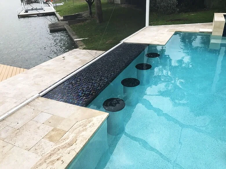 A pool edge with submerged bar stools and a dark mosaic knife-edge overflow beside a lake.