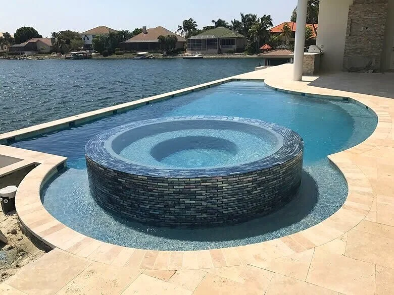 An infinity-edge pool with a round raised tiled spa in the center, overlooking a canal with waterfront homes.