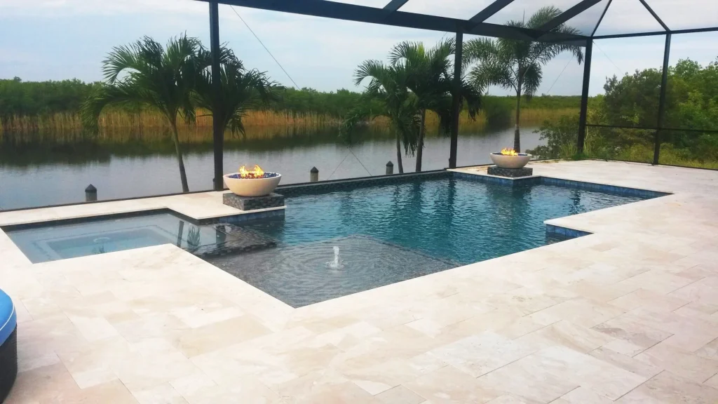 A geometric pool with an attached spa and two lit fire bowls overlooks a canal, enclosed in a screened lanai with travertine decking.