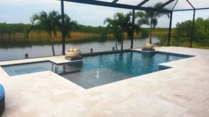 A geometric pool with an attached spa and two lit fire bowls overlooks a canal, enclosed in a screened lanai with travertine decking.