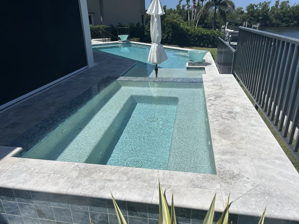A raised spa with glass tile finish sits in the foreground of a canal-front pool deck with decorative water bowls.