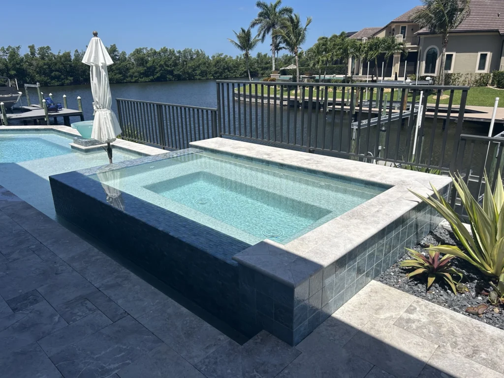 A tiled raised spa spills into a pool on a travertine deck overlooking a canal.