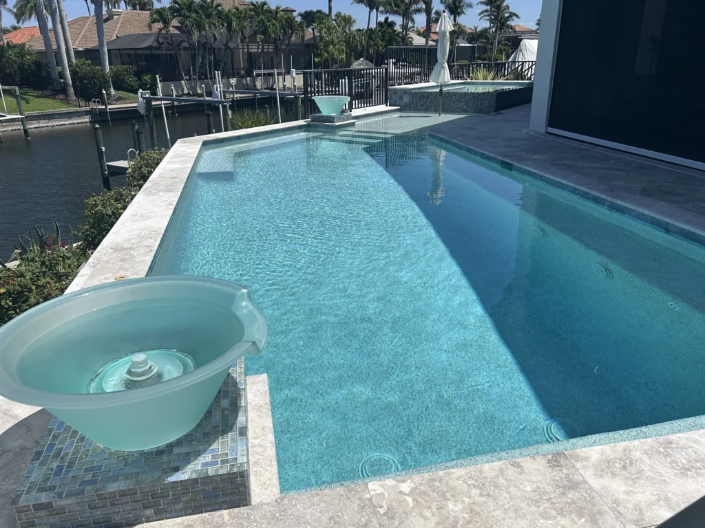 A long rectangular pool with a raised spa and water bowl fountains on a canal-front travertine deck.