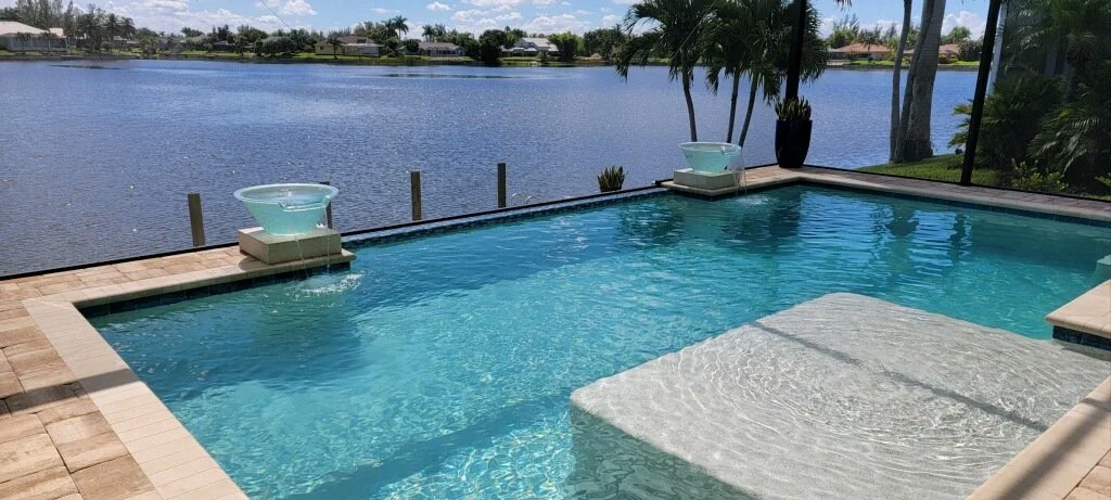 A rectangular pool with a tanning ledge and two water bowl fountains overlooks a wide canal.