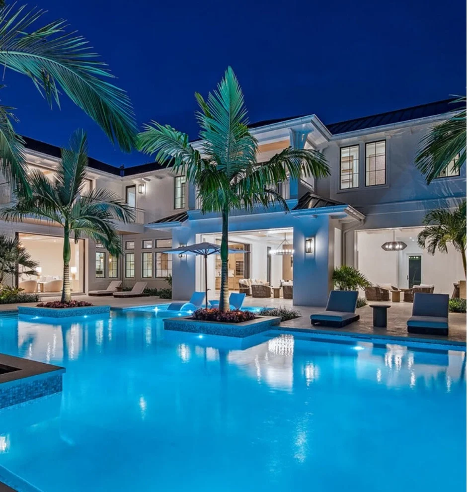 A large illuminated freeform pool at night in front of a grand two-story mansion with palm trees rising from planted islands in the water.