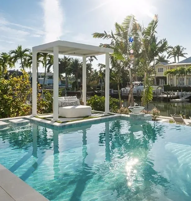 An infinity-edge pool with a shaded daybed on a sun shelf overlooking a canal lined with palm trees.