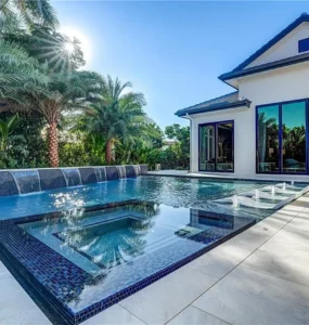 An elegant geometric pool with an all-glass-tile raised spa and a sheer descent waterfall wall beside a modern home.