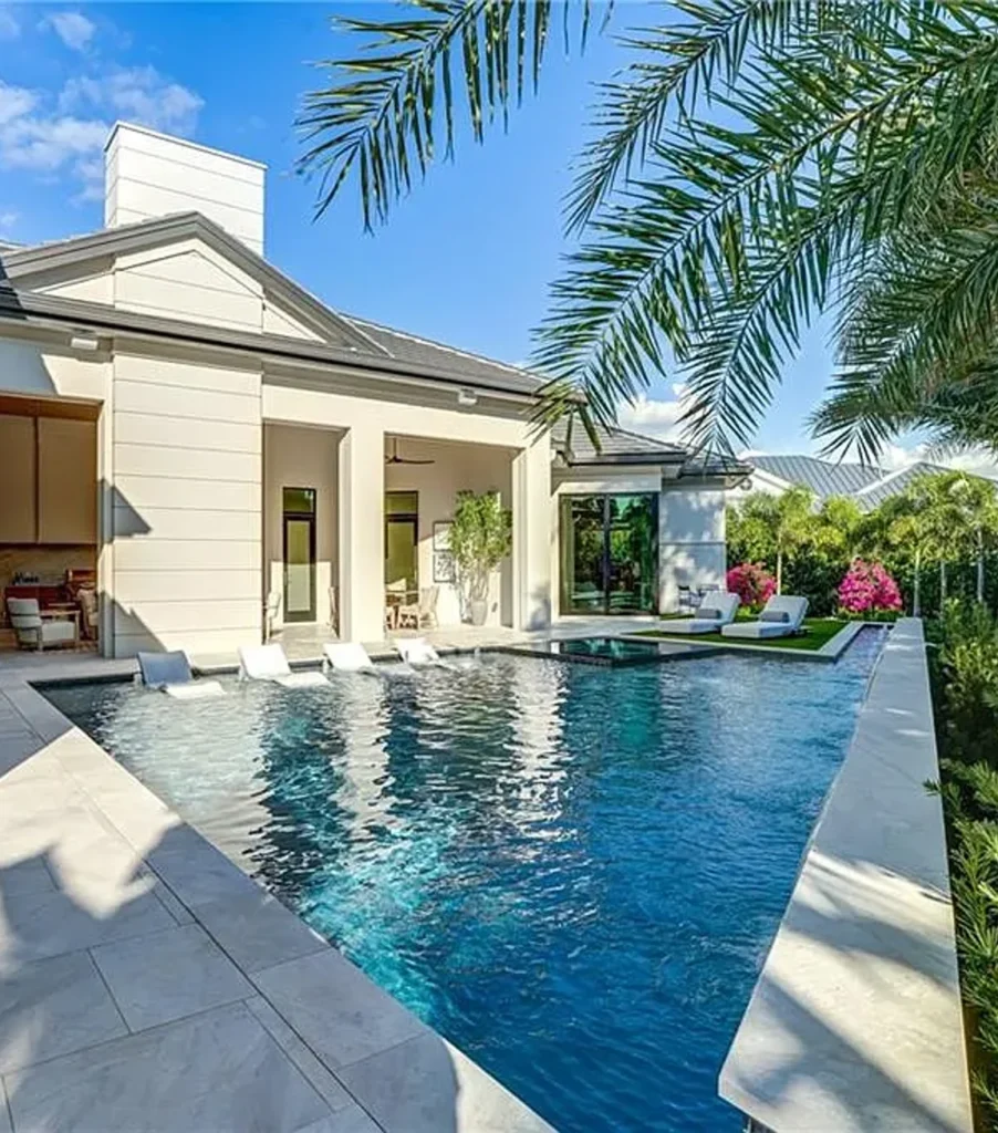 A long rectangular pool with in-water loungers runs alongside a modern home's stone patio.