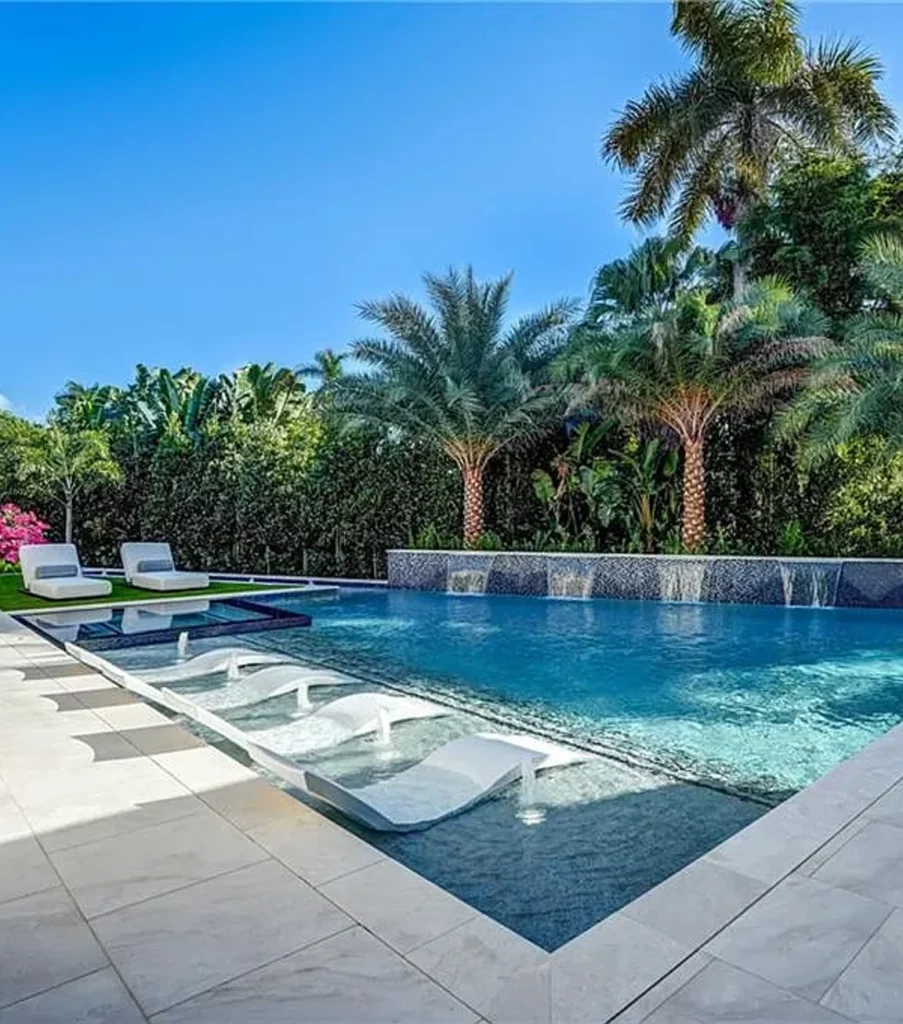 A rectangular pool with in-pool loungers and a raised mosaic tile wall with cascading water features, surrounded by lush palm trees.