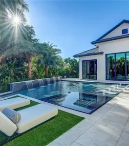 A modern rectangular pool with a raised water-wall feature and glass-edge spa sits beside a contemporary white home with turf-lined lounge areas.