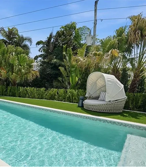 A lap pool edge with a canopied wicker daybed on artificial turf surrounded by tropical landscaping.