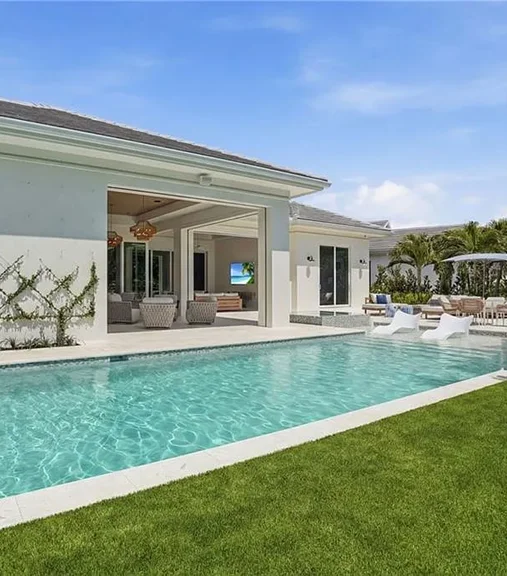 A rectangular pool with turquoise water runs alongside a modern home's open-air living space and lush lawn.
