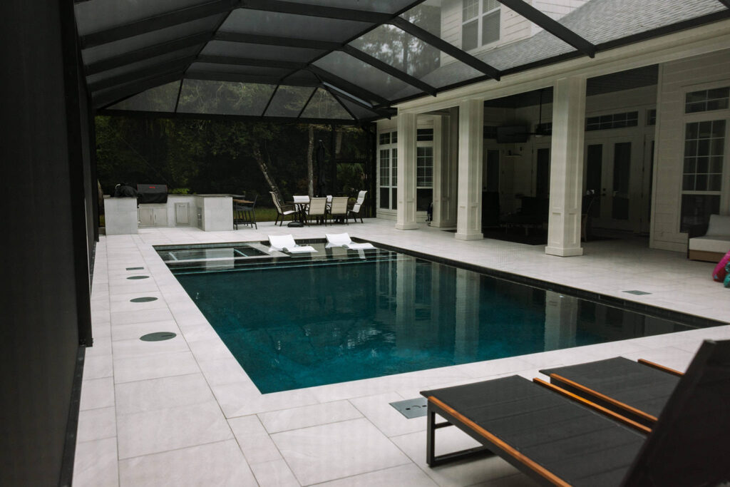 A rectangular pool with an in-pool loungers area under a screened patio enclosure attached to a large home.