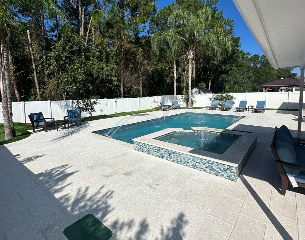 A geometric pool with a raised tiled spa and deck jets on a travertine patio surrounded by trees and a white vinyl fence.