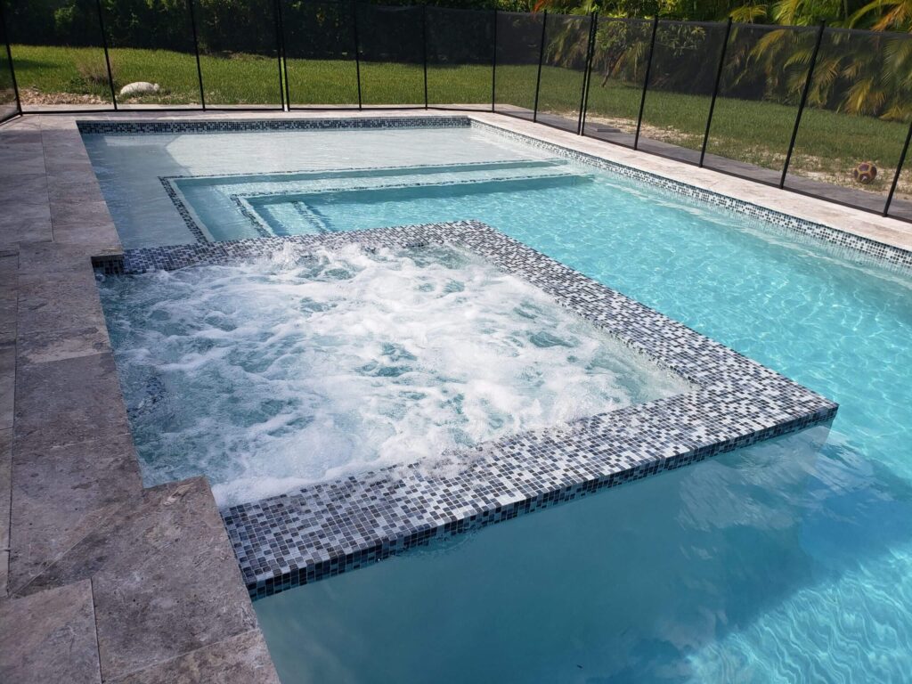 A rectangular pool with a raised mosaic-tiled spa spilling into the shallow end, surrounded by a travertine patio and safety fence.