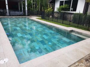A rectangular pool with textured blue-green stone tile interior and travertine deck, enclosed by a black safety fence.