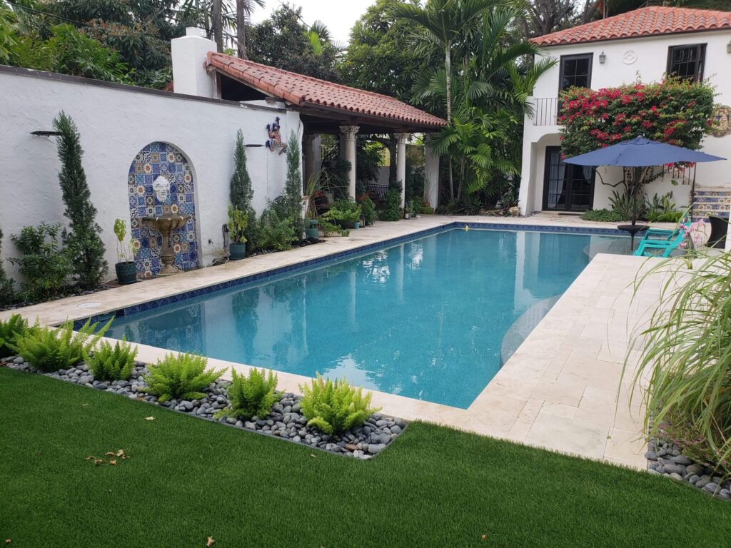 A rectangular pool with turquoise water runs alongside a Spanish-style courtyard with a tiled wall fountain and travertine decking.