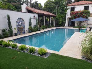 A rectangular pool with turquoise water runs alongside a Spanish-style courtyard with a tiled wall fountain and travertine decking.