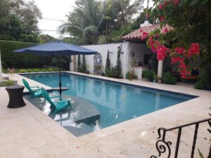A rectangular backyard pool with a submerged tanning ledge and two teal loungers under a blue umbrella, framed by a Spanish-style guest house and bougainvillea.