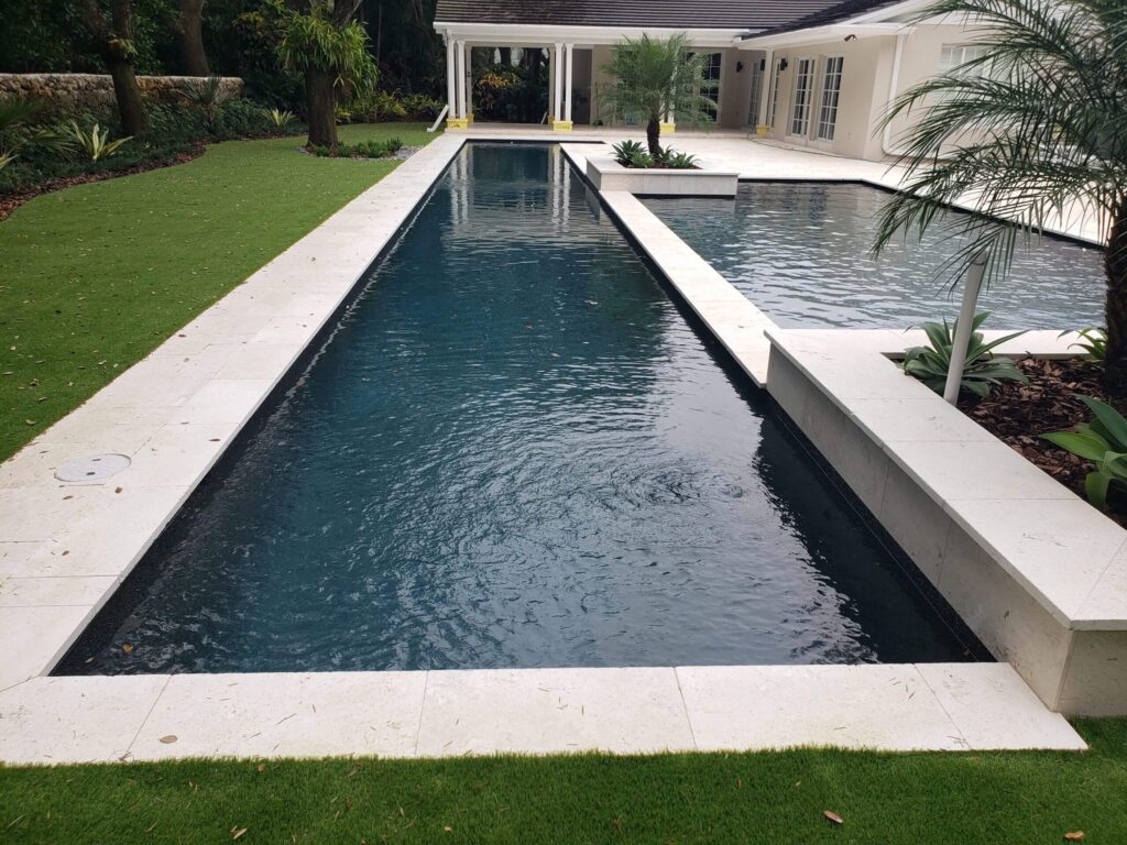 A long dark lap pool with travertine coping leads to a larger freeform pool beside a modern home.