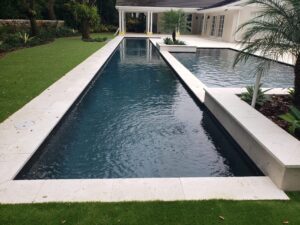 A long dark lap pool with travertine coping leads to a larger freeform pool beside a modern home.