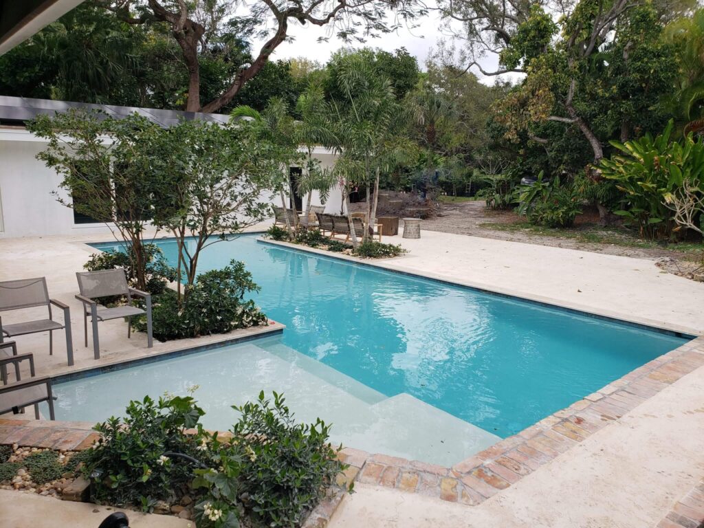 A large rectangular backyard pool with a shallow shelf corner surrounded by travertine decking and mature trees.