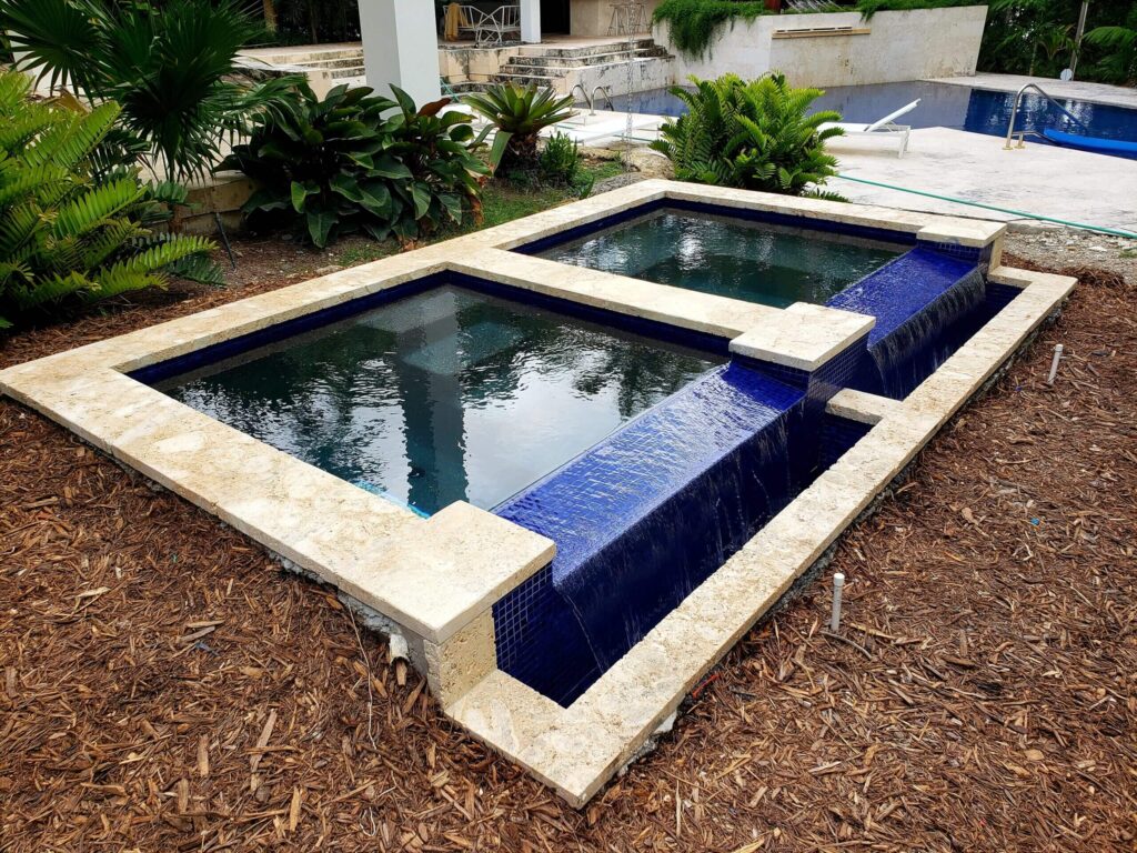 A raised two-tier water feature with blue tile spillover cascading into a basin, with a swimming pool visible in the background.