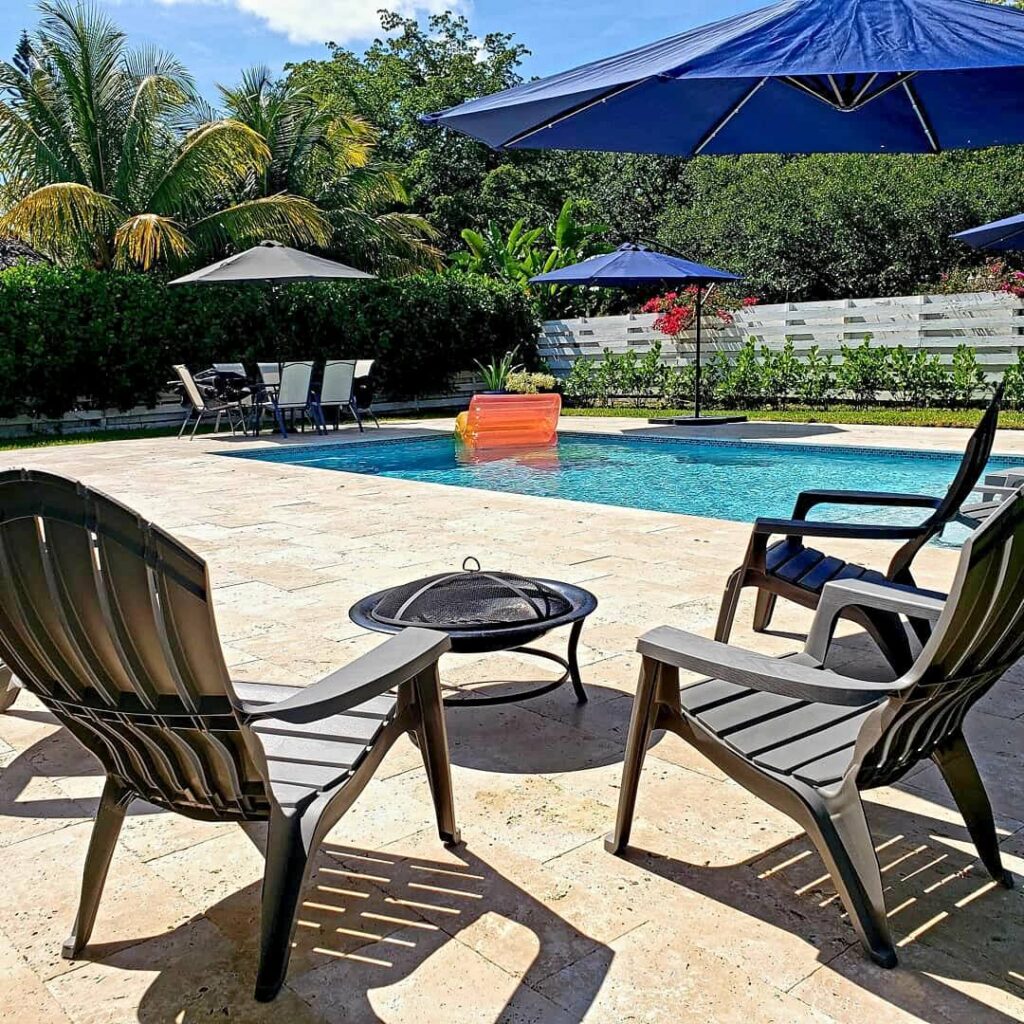 A small rectangular backyard pool with travertine deck, fire pit, and umbrellas in foreground.