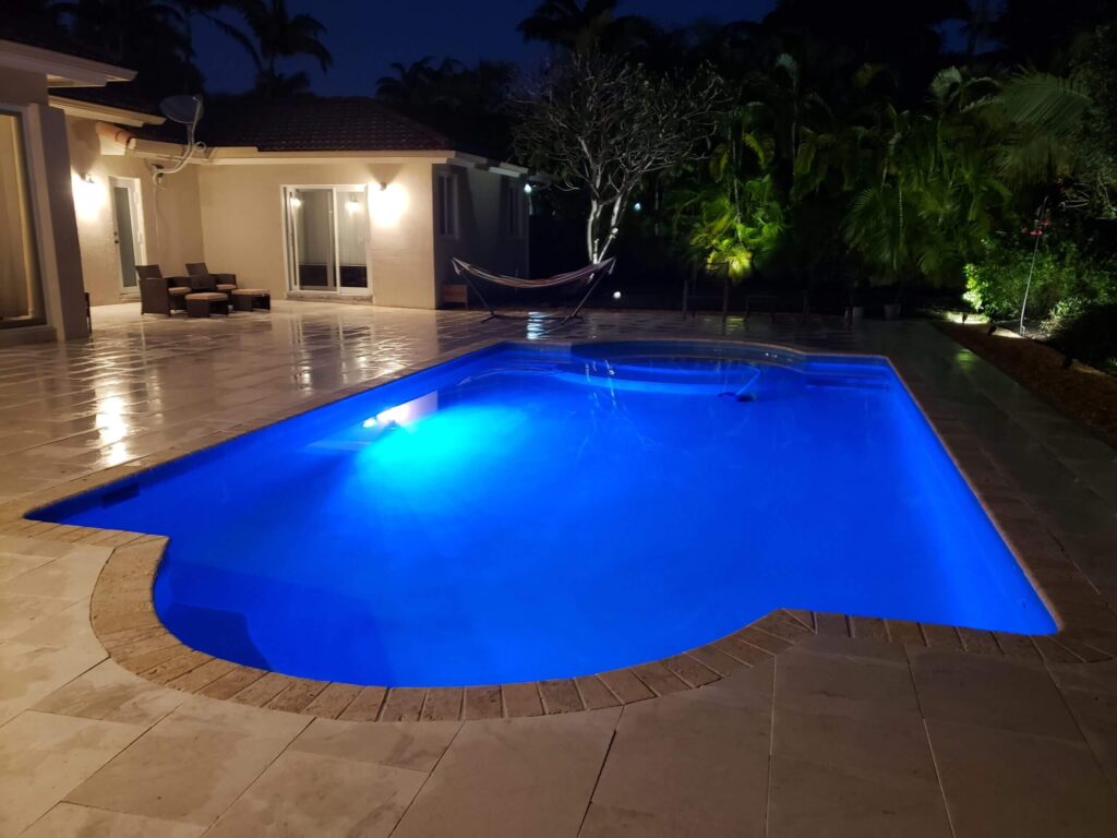 A freeform pool glowing blue at night with a raised spa and travertine deck behind a house.