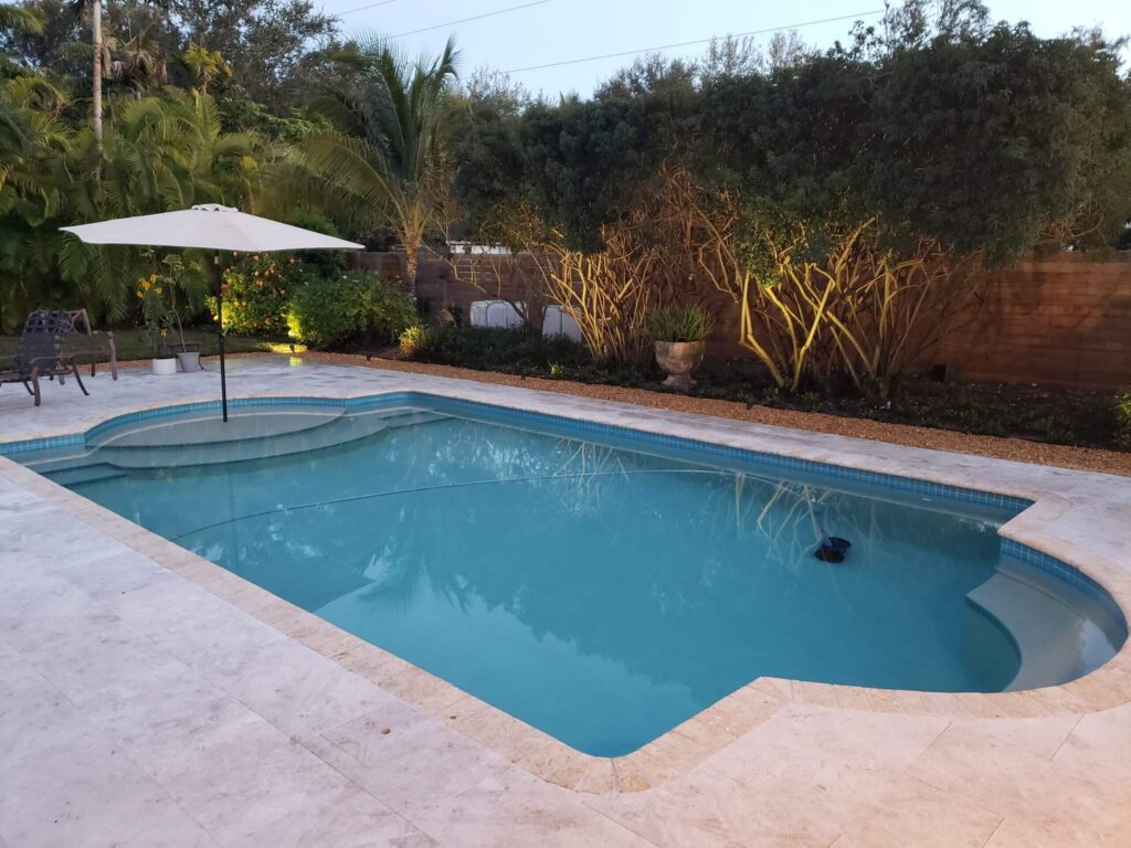 A freeform pool with a raised entry step and travertine decking in a backyard at dusk.