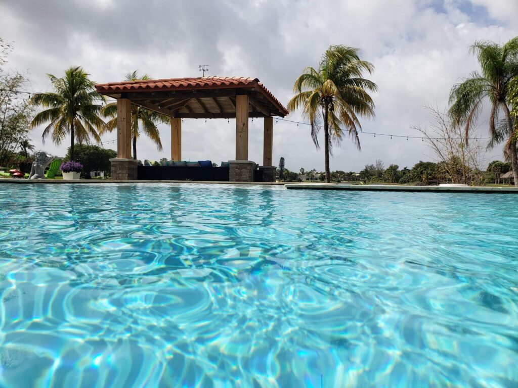Low-angle view across turquoise pool water toward a wooden pergola with a tiled roof and palm trees.