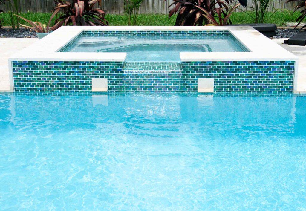 A raised spa with iridescent mosaic tile spills water into a blue pool.