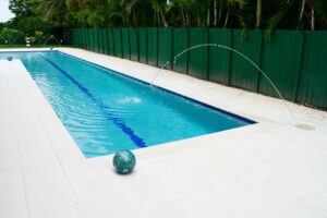A long lap pool with a blue lane marker and decorative water arc jets, bordered by a green privacy fence.