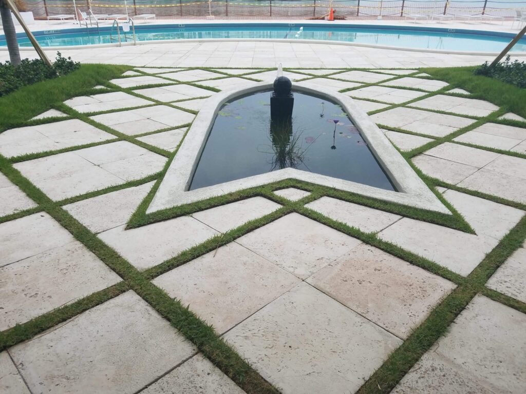 A decorative lily pond with a fountain sits in a geometric paver patio in front of a large commercial pool.